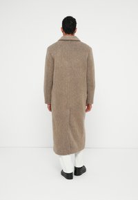 En man som står med ryggen vänd, iklädd en lång brun ulljacka, vita byxor och svarta skor mot en enkel vit bakgrund.