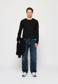 Camiseta negra de manga larga, vaqueros azules holgados y zapatillas blancas. Un suéter negro colgado sobre el brazo izquierdo. Conjunto sencillo e informal.