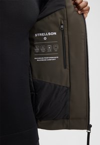 Hand öffnet den Reißverschluss der Jacke, um die Innentasche und bedruckte Etiketten zu zeigen, die wasserdicht, winddicht, leicht und komfortabel sind.