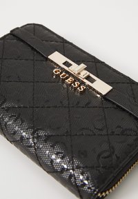Portefeuille noir texturé avec un motif matelassé, doté d'un fermoir en métal doré et du logo "GUESS" affiché en évidence sur le devant.