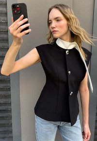 Czarna dopasowana bluzka z wysokim białym kołnierzykiem, trzema srebrnymi guzikami i dramatycznym krawatem. Dżinsy są jasnoniebieskie z lekkim odcieniem.