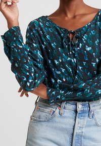 Kobieta ubrana w niebiesko-czarną bluzkę z abstrakcyjnym wzorem i zawiązanym dekoltem, w połączeniu z jasnoniebieskimi spodniami o wysokim stanie, z założonymi rękami.