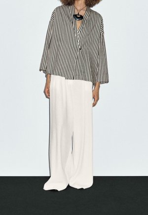 Person wearing a black-and-white vertical striped jacket, wide white pants, and a large black pendant necklace.