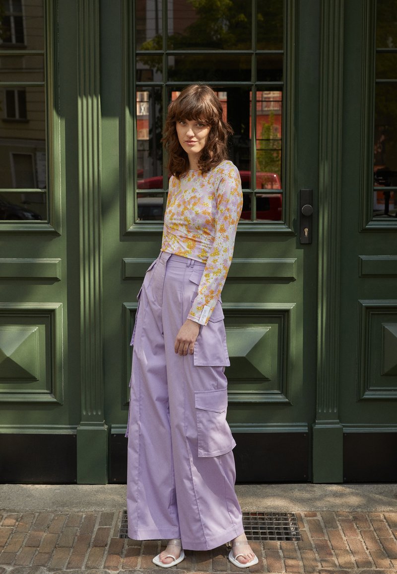 Top floral de mangas compridas em amarelo pastel e rosa, combinado com calças de carga lilás soltas, com grandes bolsos. Calçado leve, a posar ao lado de uma porta verde.