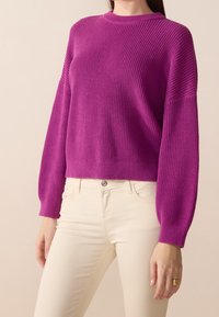 Fuksjowy sweter z dzianiny z okrągłym dekoltem i szerokimi rękawami, w połączeniu z kremowymi dżinsami. Gładka tekstura i prążkowane akcenty na rękawach oraz u dołu swetra.
