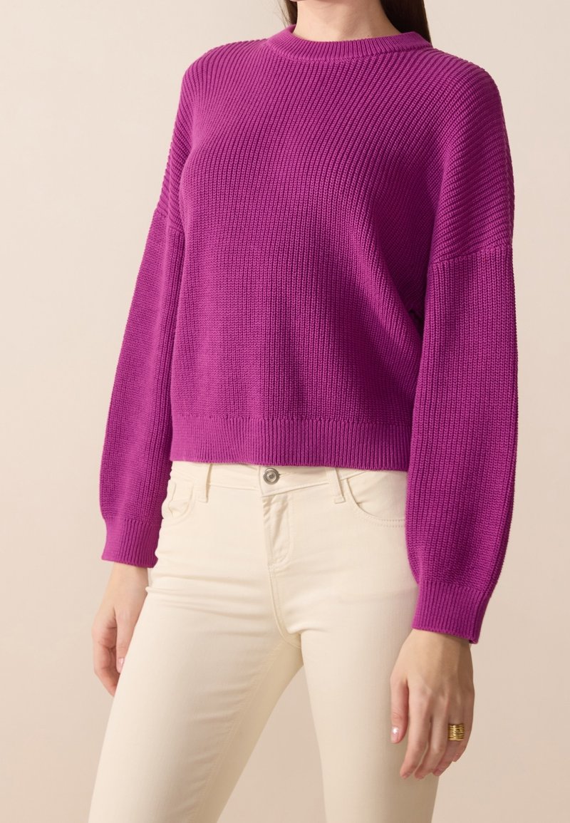 Fuksjowy sweter z dzianiny z okrągłym dekoltem i szerokimi rękawami, w połączeniu z kremowymi dżinsami. Gładka tekstura i prążkowane akcenty na rękawach oraz u dołu swetra.