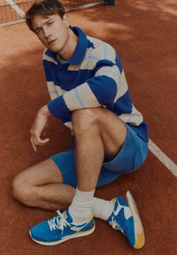 Niebieski i kremowy sweter w paski, niebieskie szorty oraz niebieskie buty sportowe z żółtymi i białymi akcentami na powierzchni kortu z czerwonej gliny.