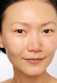 Gros plan sur un teint lisse, brun clair avec de légères imperfections, mettant en valeur des yeux marron foncé et des cheveux noirs droits. Maquillage minimal appliqué.