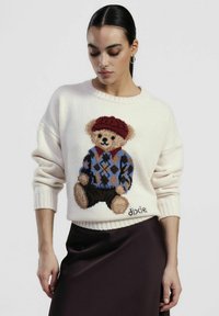 Maglione lavorato a maglia color crema con grafica di un orso con cappello rosso e maglione a rombi. Polsini e orlo a coste con dettagli "dixie". Texture semplice.