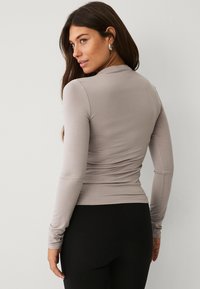 Langärmliges, eng anliegendes Oberteil aus hellgrauem Stoff, mit rundem Ausschnitt und gerafftem Detail auf der Rückseite. Kombiniert mit schwarzen Leggings.