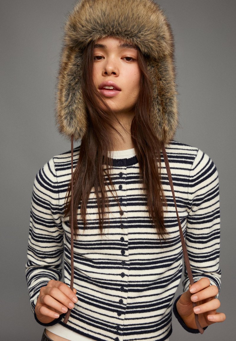 Fiatal nő csíkos, gombos pulóvert és bolyhos csuklyás sapkát visel, kezében tartja a sapka barna pántjait, szürke háttér előtt.