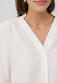 Chemisier blanc avec un col en V, fermeture à boutons et texture douce et lisse. Présente un tissu froncé aux coutures des épaules et un design classique.