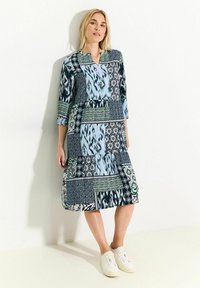 Vestido de patchwork con varios patrones en azul, verde y negro, mangas 3/4, escote en forma de V y falda escalonada, combinado con zapatillas blancas.