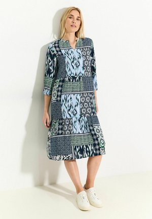 Vestito patchwork con vari pattern blu, verde e nero, maniche 3/4, scollo a V e gonna a strati, abbinato a sneakers bianche.