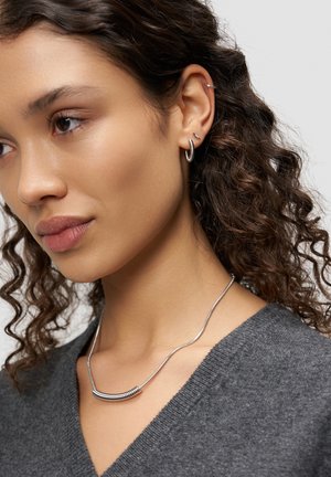 Silberne Halskette mit einem gebogenen Stabdetail, getragen über einem dunkelgrauen Pullover, ergänzt durch mehrere silberne Ohrringe im Ohr des Models.