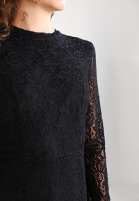 Kvinna klädd i en svart spetsklänning med långa ärmar, som visar ett blommigt spetsmönster och hög hals mot en enkel ljus bakgrund.