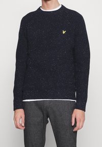 Pull texturé bleu marine avec un design côtelé, arborant un logo jaune. Le matériau comprend des taches de couleur, un col rond et des manches longues.