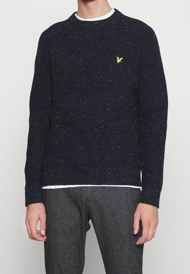 Pull texturé bleu marine avec un design côtelé, arborant un logo jaune. Le matériau comprend des taches de couleur, un col rond et des manches longues.