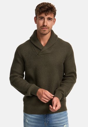 Olivegrøn strikket sweater med sjalskrave, ribbede manchetter og et struktureret mønster, parret med lyseblå denimjeans.