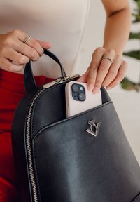 Schwarzer Leder-Rucksack mit einem Reißverschluss oben, ausgestattet mit einem silbernen Logo und einem rosa Smartphone, das teilweise in der vorderen Tasche steckt.