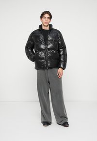 Ungdom står iført en blank svart dunjakke, løse grå bukser og svarte sko foran en enkel hvit bakgrunn.