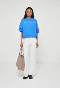 Modrá mikina s bielym logom značky, biele nohavice s voľnými nohavicami a béžová taška. Vlasy sú kučeravé a na nohách má čierne balerínky.