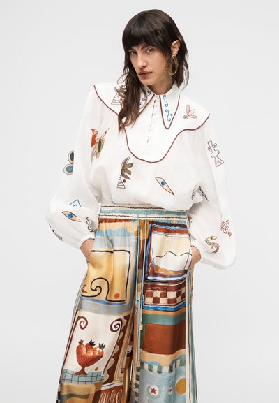 Γυναίκα που φοράει λευκό πουκάμισο με κεντημένα σχέδια και μεγάλο γιακά, σε συνδυασμό με ψηλόμεση, φαρδιά παντελόνα με πολύχρωμα αφηρημένα σχέδια.