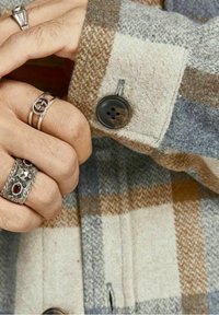 Camicia di lana a quadri in beige e grigio con un bottone scuro. Mano adornata con anelli color argento, incluso un anello decorativo con una gemma.