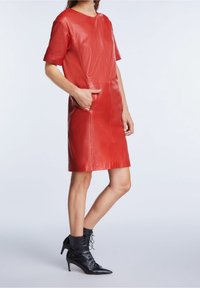 Robe en cuir rouge à manches courtes, dotée de deux poches avant et d'un col rond. Portée avec des bottines noires à lacets.