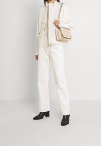 Kremowa kamizelka puchowa z wysokim kołnierzem, nałożona na jasny sweter i białe proste spodnie; zestawiona z jasną torebką i czarnymi botkami na obcasie.