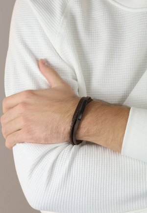 Braided brown leather bracelet with a black metal accent, worn on a wrist covered by a textured white long-sleeve shirt.