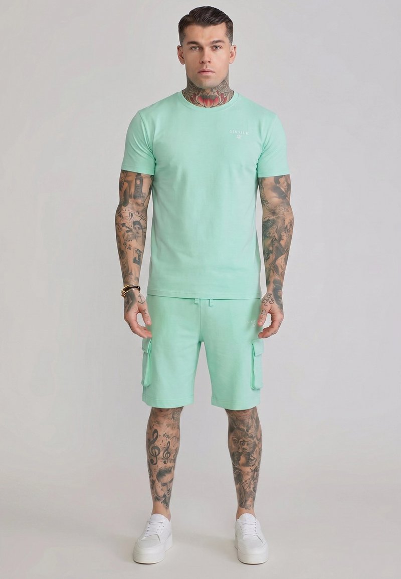 Man met volledige mouw tattoos en nektattoo, draagt een muntgroen T-shirt en cargoshorts met witte sneakers, staand tegen een effen achtergrond.