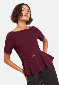 Burgundská peplum blůza s krátkými rukávy, s úzkým pasem zvýrazněným detailem stříbrné spony. Hladká textura látky, strukturovaný design.