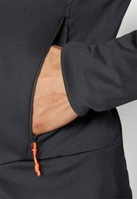 Svart treningsoverdel med en glidelåslomme og oransje snor. Stoffet er glatt og tettsittende, noe som fremhever et stilig design.