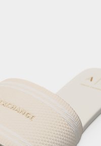 Sandale à glissière beige avec sangle texturée portant le texte embossé « EXCHANGE » et semelle intérieure lisse avec logo partiel de la marque.