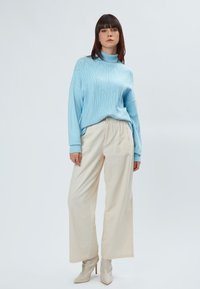 Maglione a maglia testurizzata blu chiaro con collo alto abbinato a pantaloni larghi color crema. Il modello indossa stivali alla caviglia beige lucidi.