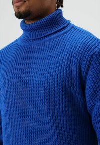 Blauwe gebreide coltrui met een geribbelde textuur en dikke stof, met een nauwsluitend ontwerp en een opvallende kraag.