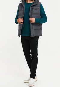 Kamizelka puffer w ciemnoszarym kolorze z kapturem, zapinana na zamek, z wieloma kieszeniami, w parze z bluza w kolorze teal i czarnymi spodniami. Białe sneakersy.