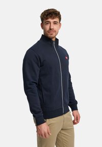 Marinblå zip-up jacka med hög krage, med en röd logotyp på bröstet och ribbade muddar och fåll för en tät passform. Mjuk tygstruktur.