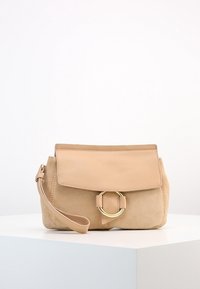 Sac à main en daim beige avec un rabat en cuir lisse, un accent circulaire doré sur le devant, et une bandoulière amovible.