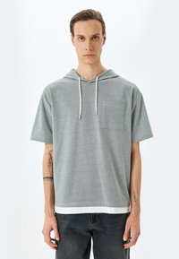 Sudadera corta de manga corta gris con un bolsillo frontal, cordones blancos y un dobladillo blanco en contraste, hecha de un tejido suave y texturizado.