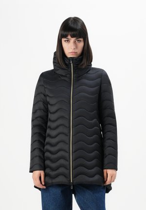 Vrouw in een zwart gewatteerd pufferjack met een hoge kraag en een ritssluiting aan de voorzijde, gecombineerd met blauwe jeans, staand tegen een witte achtergrond.