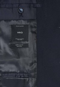 Intérieur de veste de costume bleu marine foncé montrant l'étiquette de la marque "MNG", taille EUR 50 UK USA MEX 40, et l'étiquette "Tissu stretch coupe super slim".