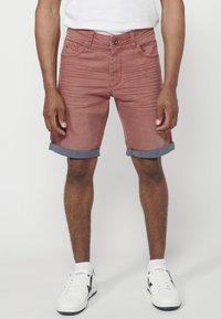 Shorts en denim rouge avec des ourlets bleus retroussés, poches avant et décoloration subtile, associés à des baskets blanches et des chaussettes mi-mollet.
