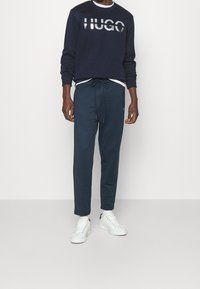 Uomo che indossa un felpa Hugo blu navy, pantaloni della tuta blu navy abbinati, una maglietta bianca sotto e sneakers bianche, in piedi.