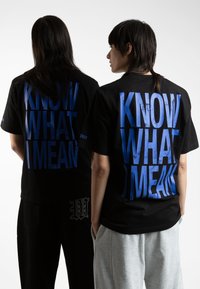To personer står ryg mod ryg iført sorte T-shirts med teksten "VED DU HVAD JEG MENER" trykt i blå. De har forskellige frisurer og afslappet tøj.