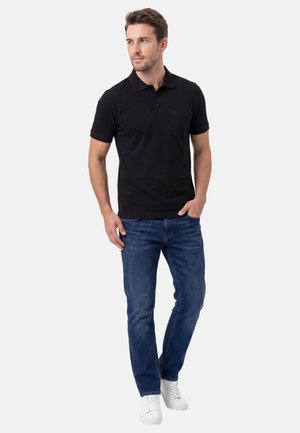 Homme portant un polo noir, un jean bleu et des baskets blanches, debout avec une main dans la poche, regardant sur le côté.