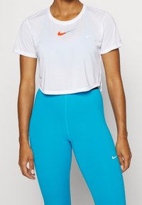 Camiseta corta blanca con logos de Nike en naranja y azul. Tela ligera y transpirable. Combinada con leggings ajustados de un brillante color azul.