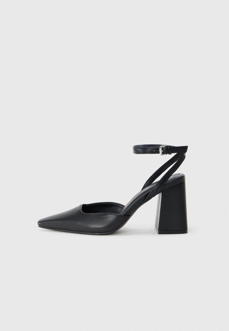 Black leather pointy-toe pump with an ankle strap and a chunky heel. Flat texture, sleek design, and minimalistic aesthetics.