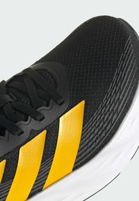 Close-up of black athletic shoe with yellow stripes, black laces, and white sole, highlighting breathable mesh and textured design details.